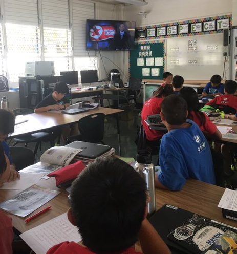 Students in classroom watching newscast on overhead TV monitor