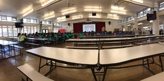 Wide angle view of cafeteria tables with student group in green shirts close to the stage