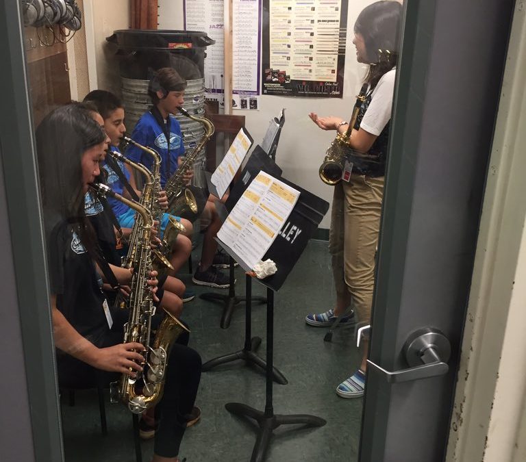 view through door window woman standing and 4 seated students playing the saxophone