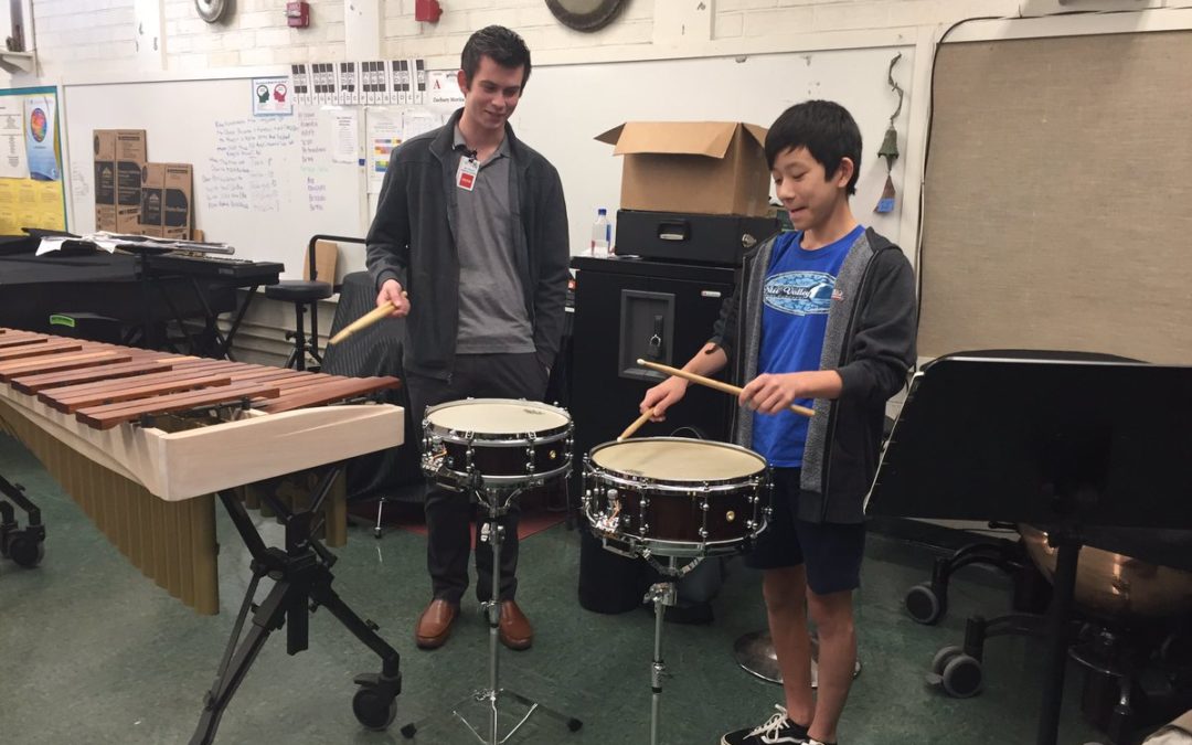 Man in black jacket watching boy playing snare drum in music room