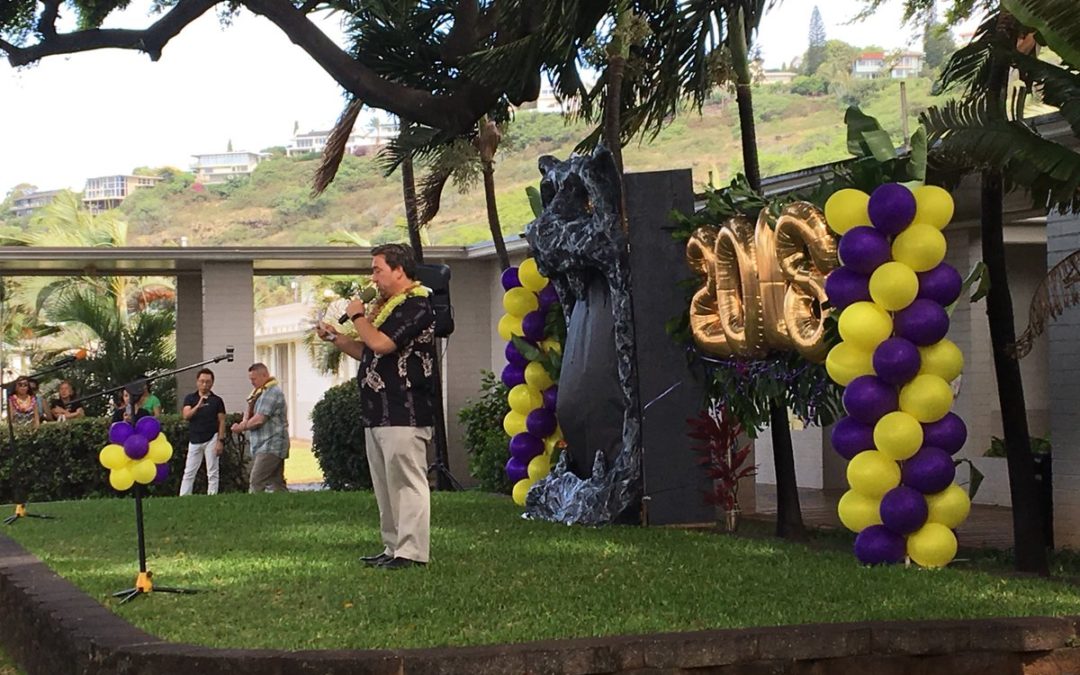 Man with lei speaking on grass stage with purple, yellow, and 2018 balloons