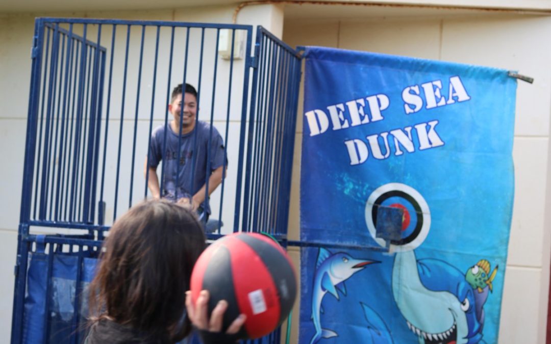 Teacher sits in dunk book with student holding black and red ball ready to throw, target with deep sea dunk sign